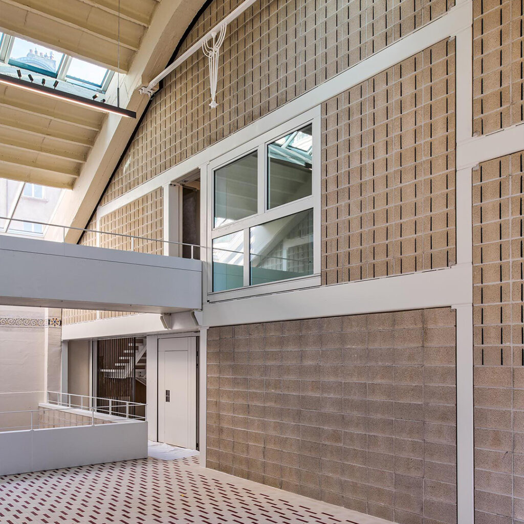 A modern indoor architectural space featuring a high ceiling, skylights, grey brick walls, a window, and a white door. The floor has a red and white geometric tile pattern.