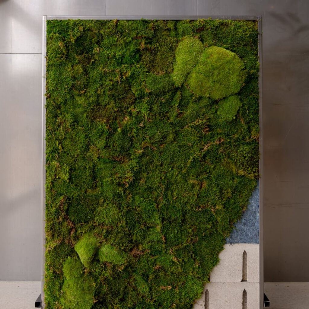 A large, vertical green moss wall installation with geometric concrete elements at the bottom, mounted on a plain metallic backdrop.