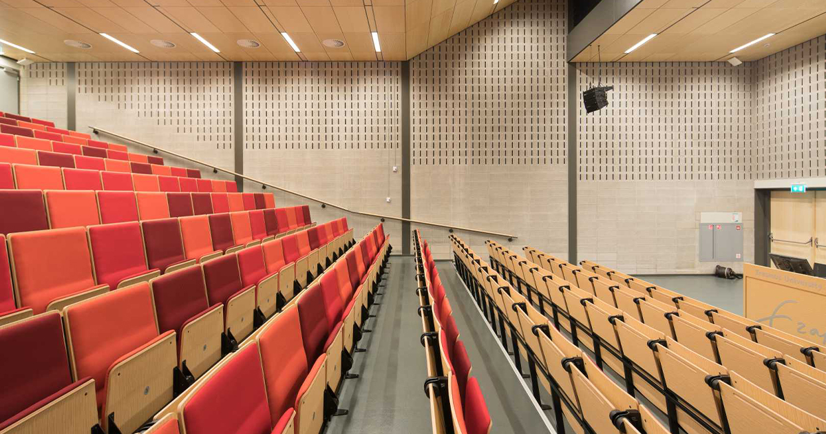 Empty lecture hall with tiered red and orange seating, grey floors, beige brick walls, and a wooden ceiling with spotlights.